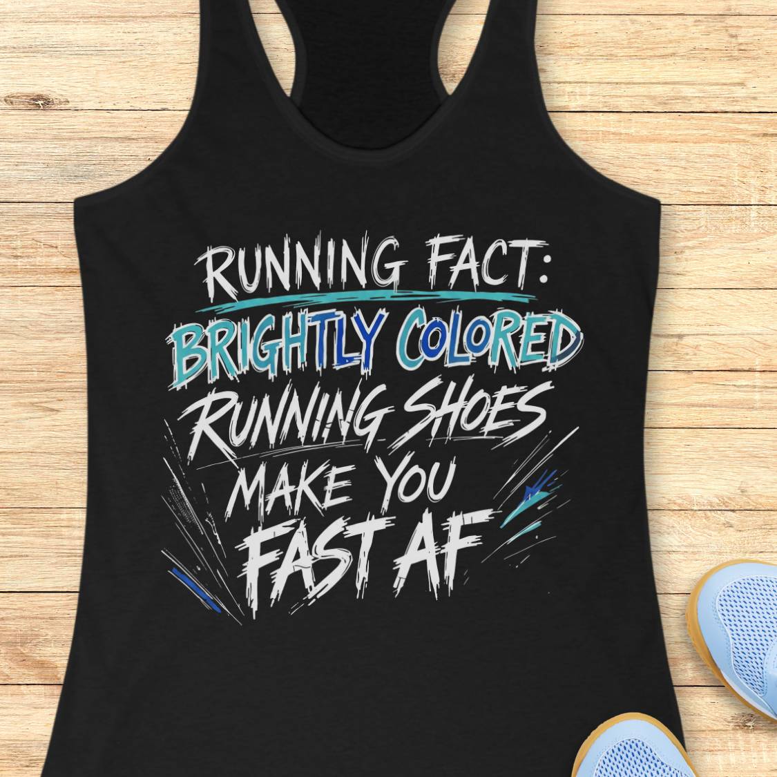 A black racerback tank top laid flat on a light wood surface, featuring a bold graphic text print that reads 'RUNNING FACT: BRIGHTLY COLORED RUNNING SHOES MAKE YOU FAST AF' in white, blue, and teal lettering. Partial light blue sneakers are visible at the bottom right edge.