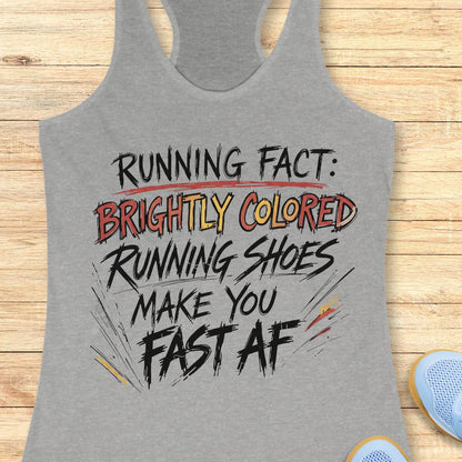A heather gray racerback tank top with a colorful graphic text print reading 'RUNNING FACT: BRIGHTLY COLORED RUNNING SHOES MAKE YOU FAST AF', displayed flat on a light wood background with partial blue sneakers visible at the bottom right.
