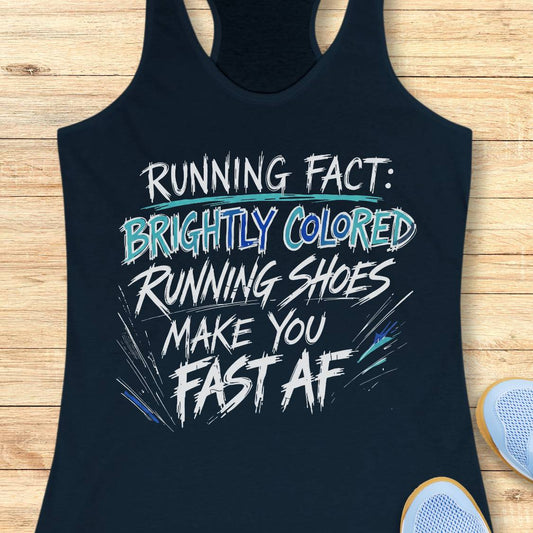 A navy racerback tank top laid flat on a light wood background, featuring a bold white and light blue graphic text that reads 'RUNNING FACT: BRIGHTLY COLORED RUNNING SHOES MAKE YOU FAST AF'. Partial white sneakers are visible at the bottom right as styling props.
