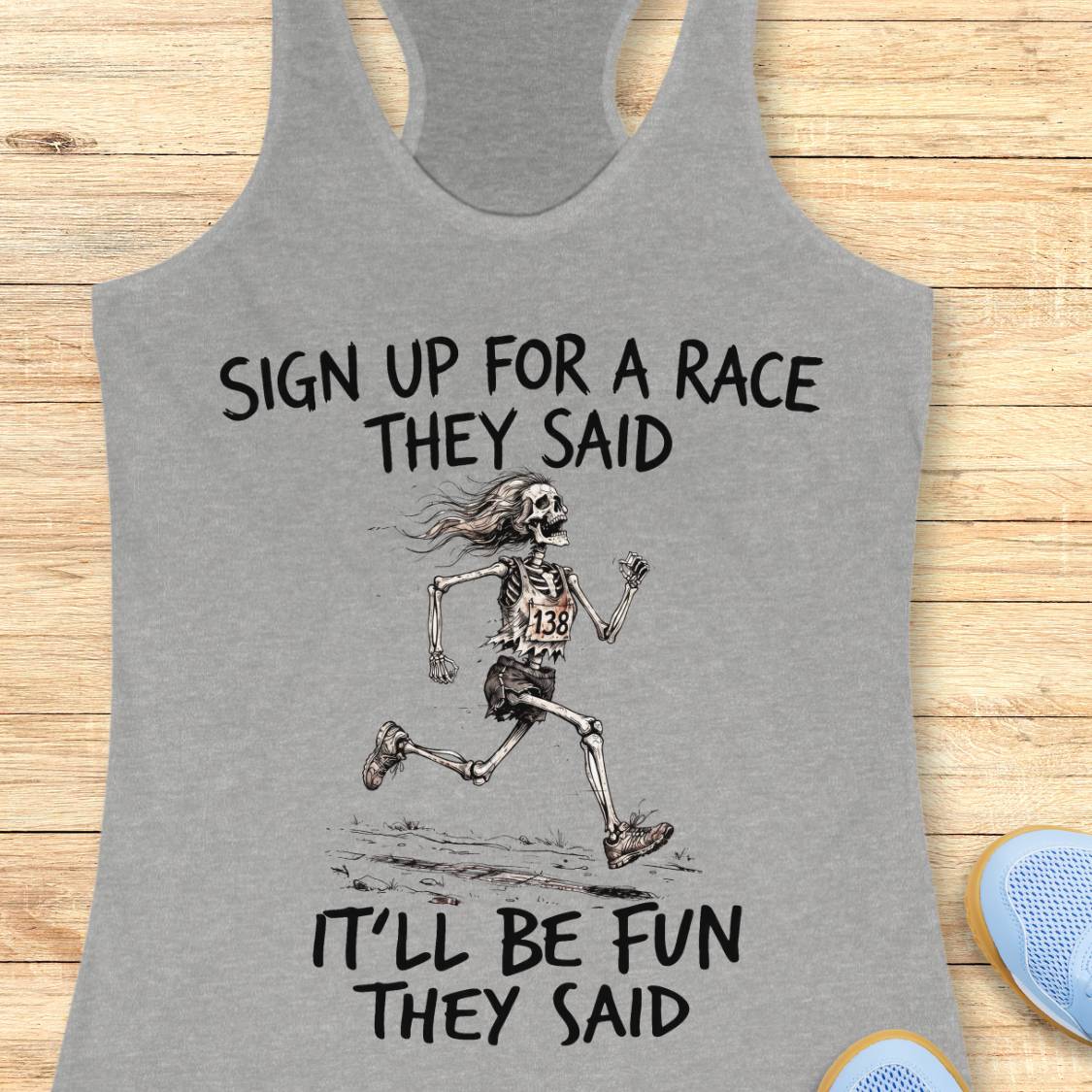 A heather gray racerback tank top with a black graphic of a skeleton running and the text 'SIGN UP FOR A RACE THEY SAID IT'LL BE FUN THEY SAID', shown on a light wood background with partial blue shoes visible at the edges.