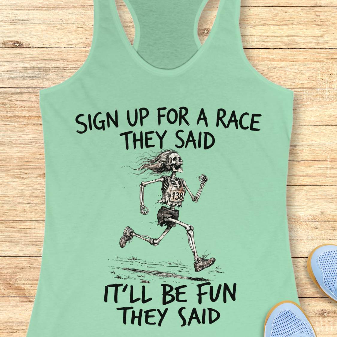 A mint green racerback tank top laid flat on a light wood background, featuring a black graphic of a running skeleton and the text 'SIGN UP FOR A RACE THEY SAID IT'LL BE FUN THEY SAID.' Partial blue shoes are visible at the bottom corners as styling props.