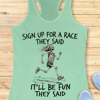 A mint green racerback tank top laid flat on a light wood background, featuring a black graphic of a running skeleton and the text 'SIGN UP FOR A RACE THEY SAID IT'LL BE FUN THEY SAID.' Partial blue shoes are visible at the bottom corners as styling props.