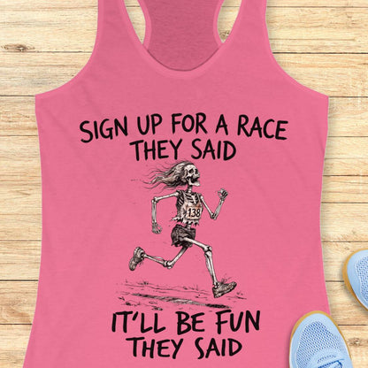 A pink racerback tank top laid flat on a light wood surface, featuring a black graphic of a running skeleton and the text 'SIGN UP FOR A RACE THEY SAID IT'LL BE FUN THEY SAID.' Partial blue shoes are visible at the lower right as styling props.