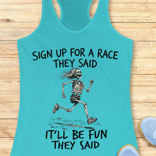 A turquoise racerback tank top laid flat on a light wood surface, featuring a black graphic of a running skeleton and the text 'SIGN UP FOR A RACE THEY SAID IT'LL BE FUN THEY SAID.' A partial blue shoe is visible at the lower right as a styling prop.