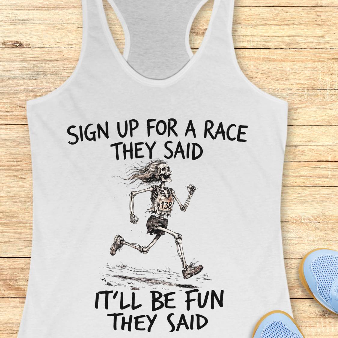 A white racerback tank top laid flat on a light wood surface, printed with the phrase 'SIGN UP FOR A RACE THEY SAID IT'LL BE FUN THEY SAID' around a sketch of a running skeleton wearing a race bib. Part of a blue shoe is visible at the lower right as a styling prop.