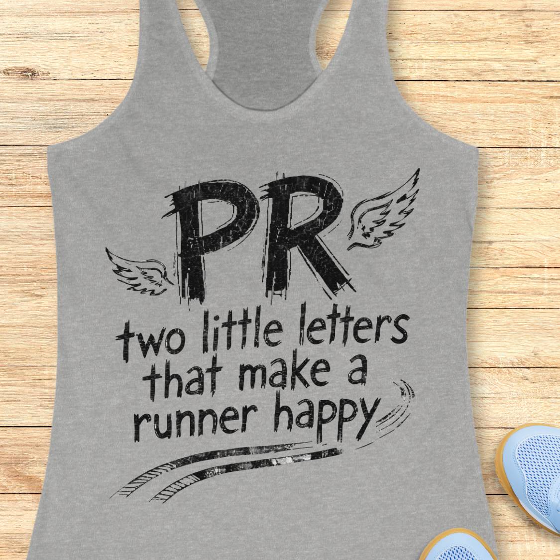 A heather gray racerback tank top with a black graphic print reading 'PR two little letters that make a runner happy' with wings and track lines, shown on a light wood background with partial blue shoes at the corner.
