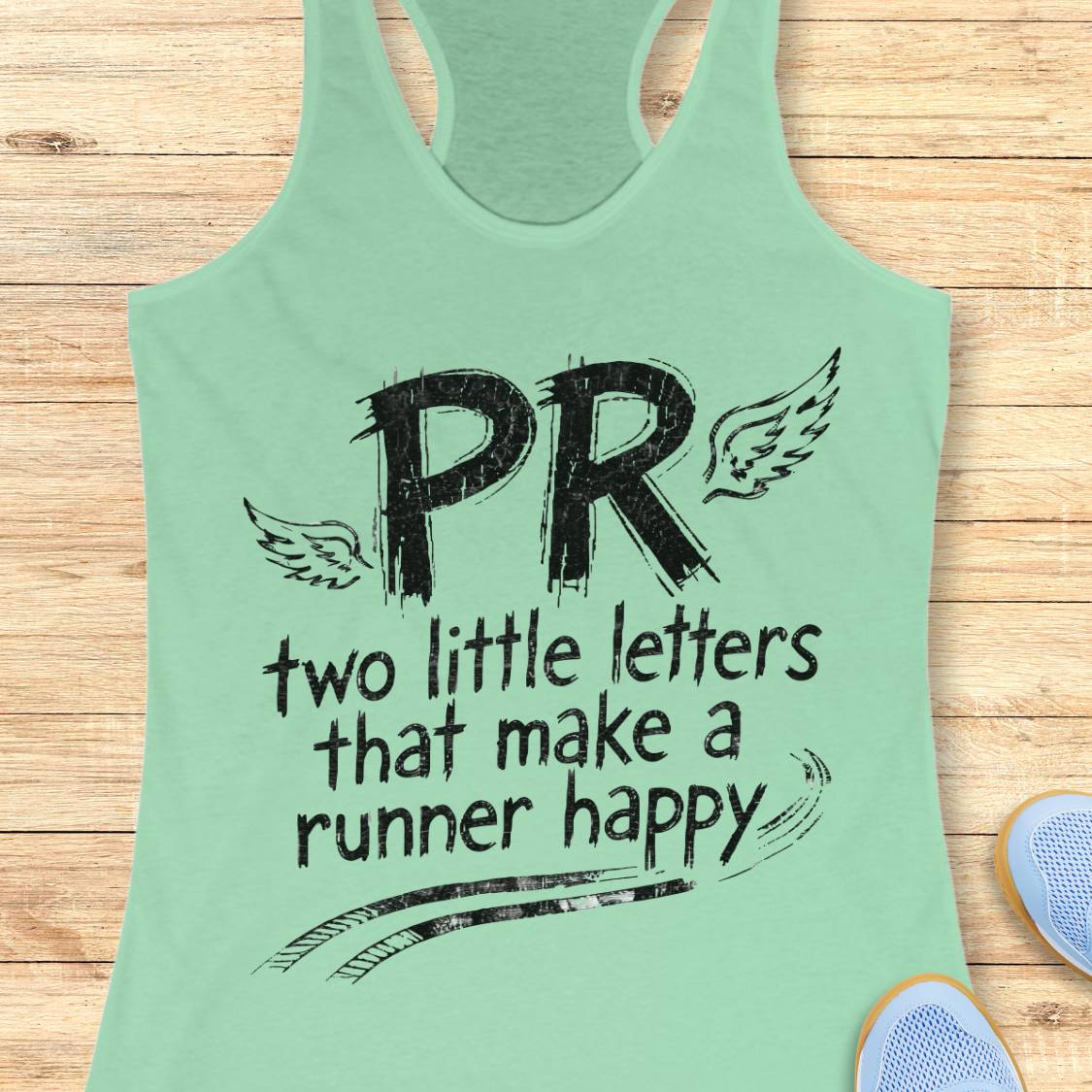 A mint green racerback tank top with a black graphic print reading 'PR two little letters that make a runner happy' with wings and track strip marks, laid flat on a light wood background with partial shoes visible at the edges.
