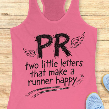 A pink racerback tank top laid flat on a light wood surface, featuring black distressed text that reads 'PR two little letters that make a runner happy' with wing graphics. Partial blue sneakers are visible at the edges as styling props.