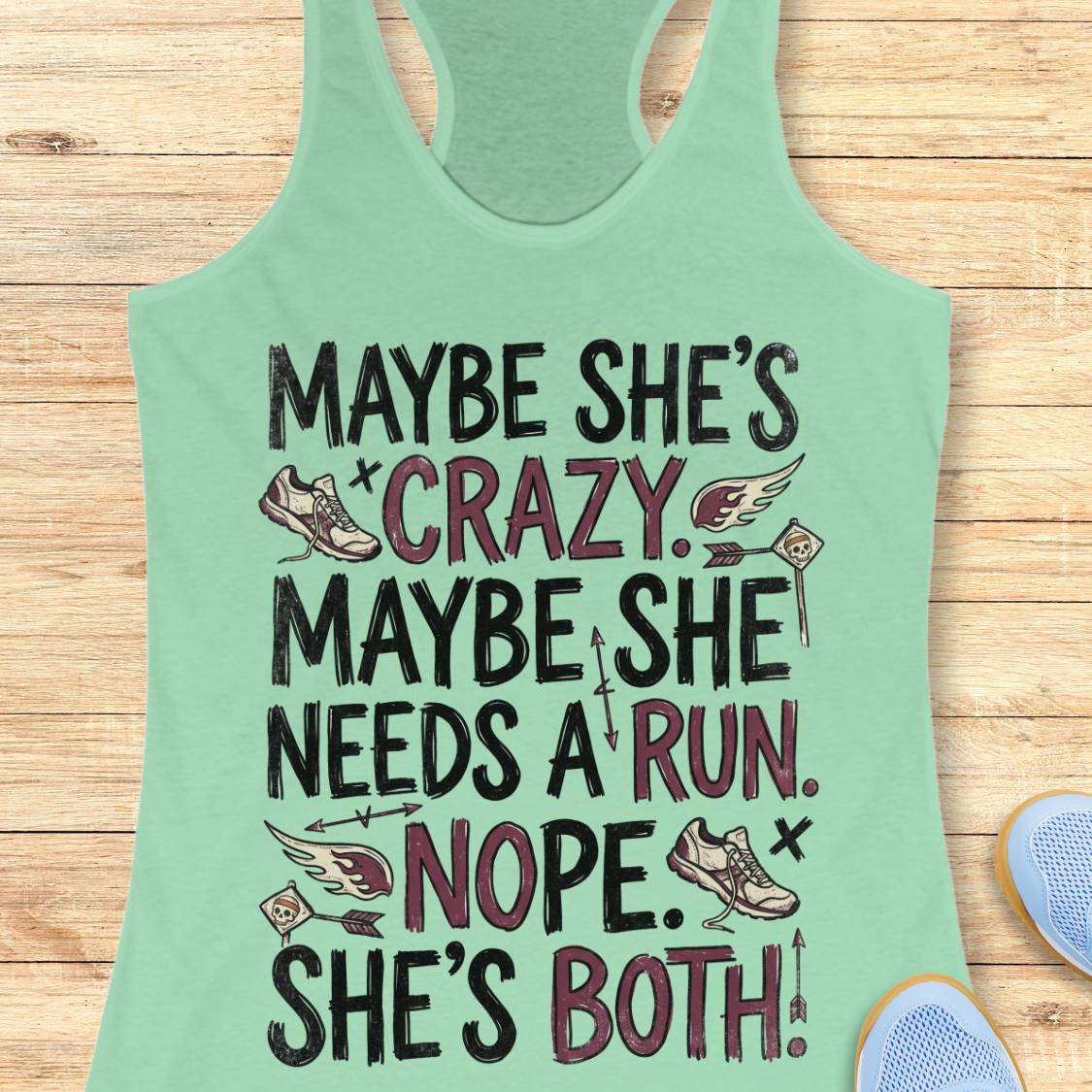 A mint green racerback tank top with a humorous graphic print, laid flat on a light wood background. The graphic features the text 'MAYBE SHE'S CRAZY.