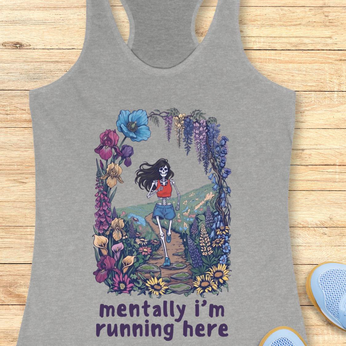 A heather gray racerback tank top laid flat on a light wood background, featuring a colorful graphic of a skeleton woman running through a flower arch, with the text 'mentally i'm running here' printed below.