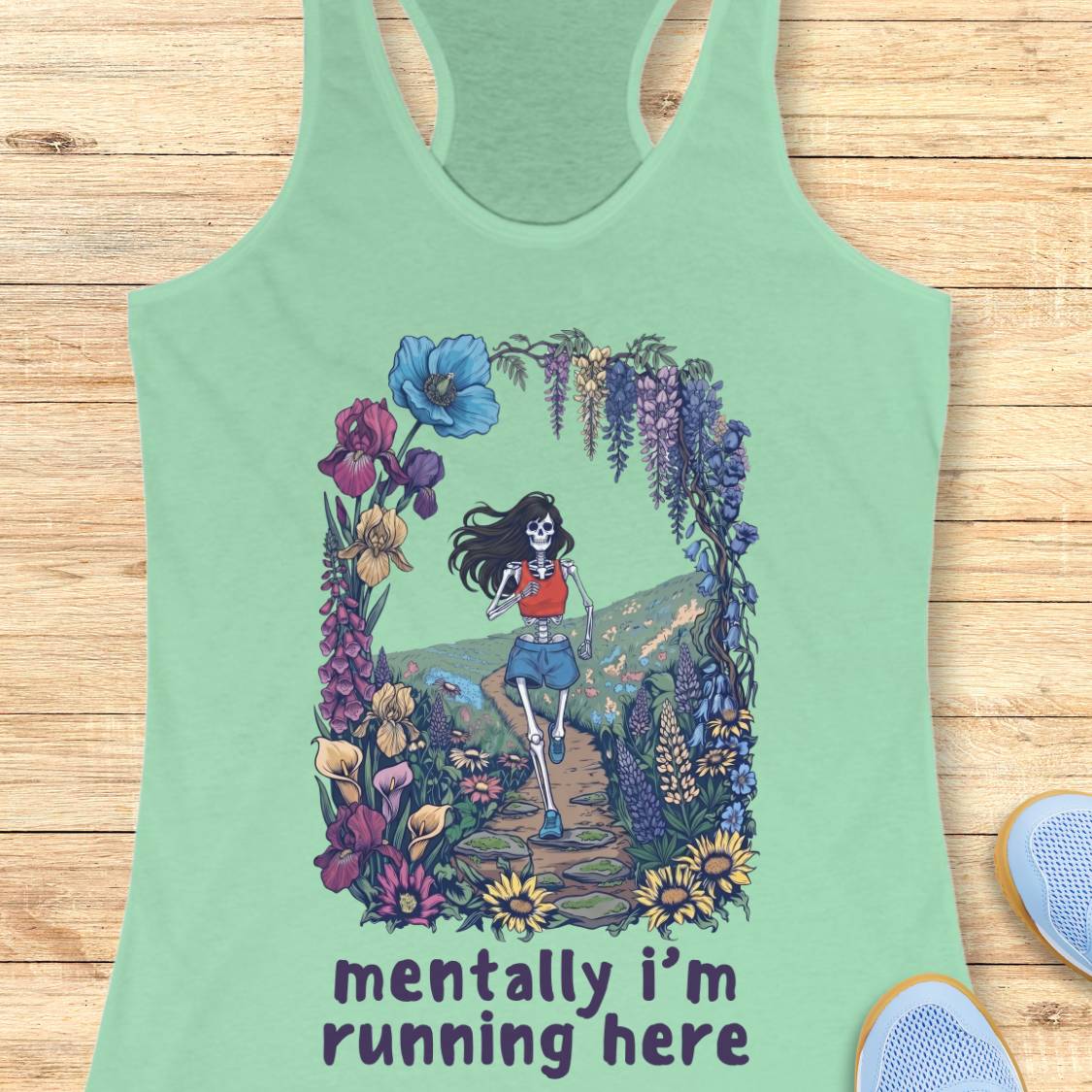 A mint green racerback tank top laid flat on a light wood background, featuring a colorful graphic of a skeleton runner framed by wildflowers and the text 'mentally i'm running here' printed below.