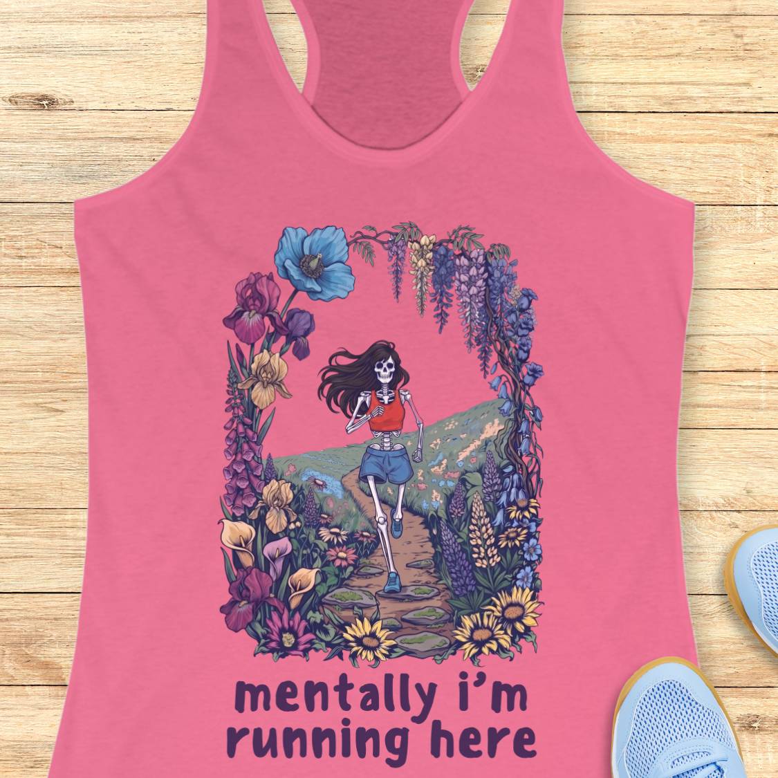 A pink racerback tank top laid flat on a light wood surface, featuring a colorful graphic of a skeleton runner framed by flowers and the text 'mentally i'm running here'. Partial blue sneakers are visible at the lower right as styling props.