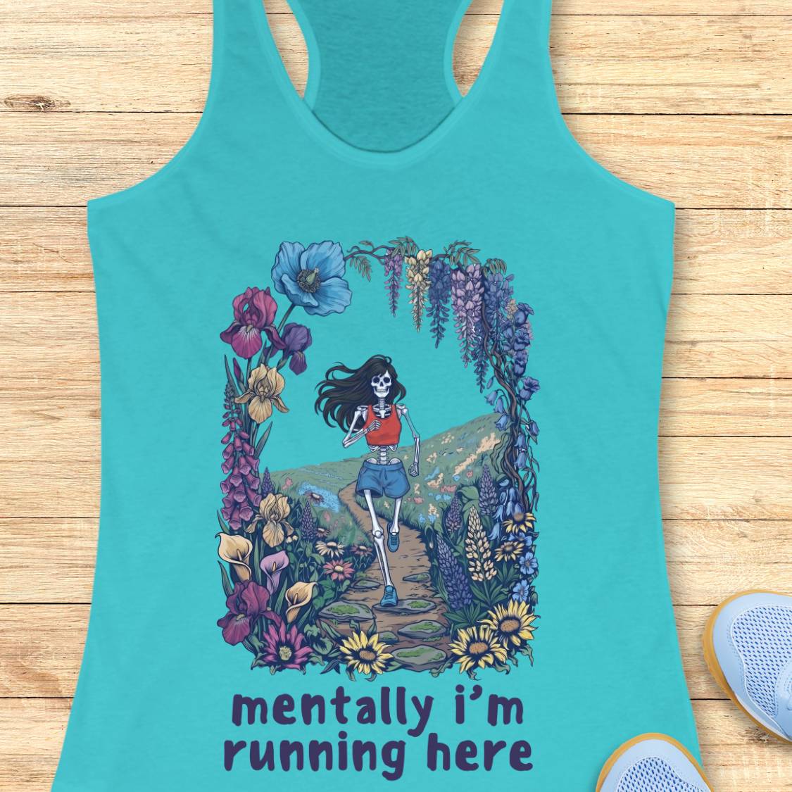 A turquoise racerback tank top laid flat on a light wood surface, featuring a colorful graphic of a running skeleton framed by flowers, with the text 'mentally i'm running here' printed below.