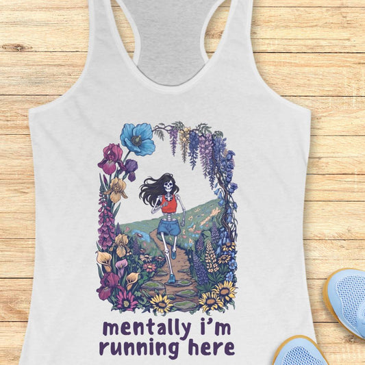 A white racerback tank top laid flat on a light wood background, featuring a colorful graphic of a woman running through flowers with the text 'mentally i'm running here'. A pair of blue sneakers is partially visible at the bottom right.