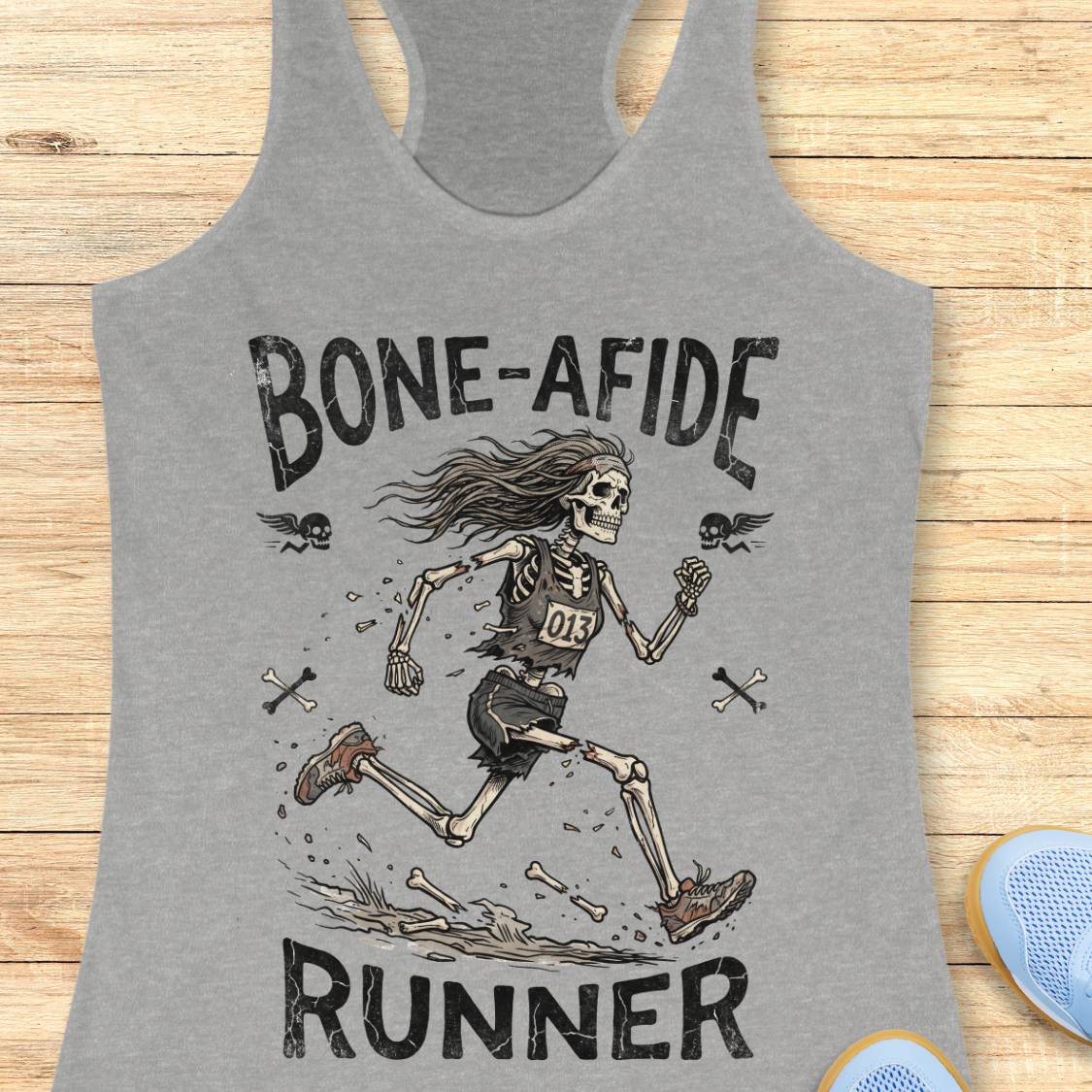 A heather gray racerback tank top laid flat on a light wood surface, featuring a distressed black graphic of a running skeleton with the text 'BONE-AFIDE RUNNER' and small skull-and-crossbones details.