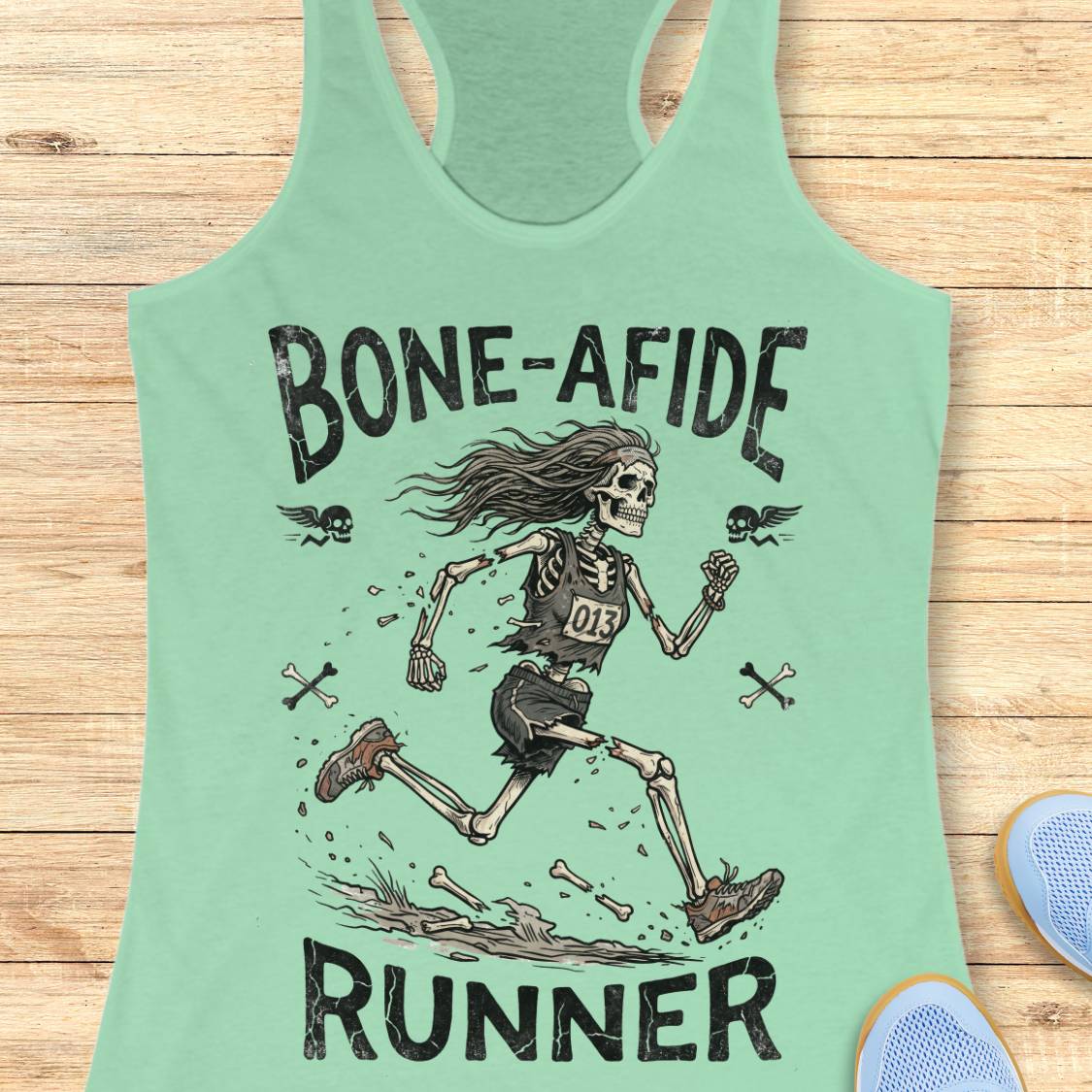 A mint green racerback tank top laid flat on a light wood background, featuring a distressed black graphic of a running skeleton with the text 'BONE-AFIDE RUNNER' and small flying bones around it.