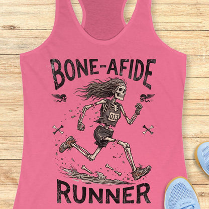 A pink racerback tank top laid flat on a light wood surface, featuring a large black graphic of a running skeleton with the words 'BONE-AFIDE' above and 'RUNNER' below. Partial blue shoes are visible at the edges as styling props.