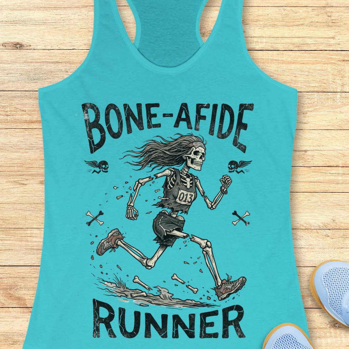 A turquoise racerback tank top laid flat on a light wood surface, featuring a large graphic of a running skeleton with the words 'BONE-AFIDE RUNNER' and small bone illustrations. Part of a blue shoe is visible at the lower right edge.