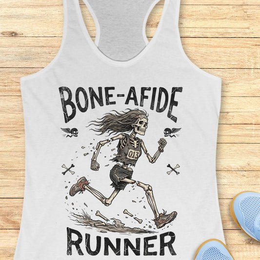A white racerback tank top laid flat on a light wood surface, featuring a black graphic of a running skeleton with the text 'BONE-AFIDE RUNNER' and small skull-and-crossbones details. Part of a blue shoe is visible at the lower right edge.