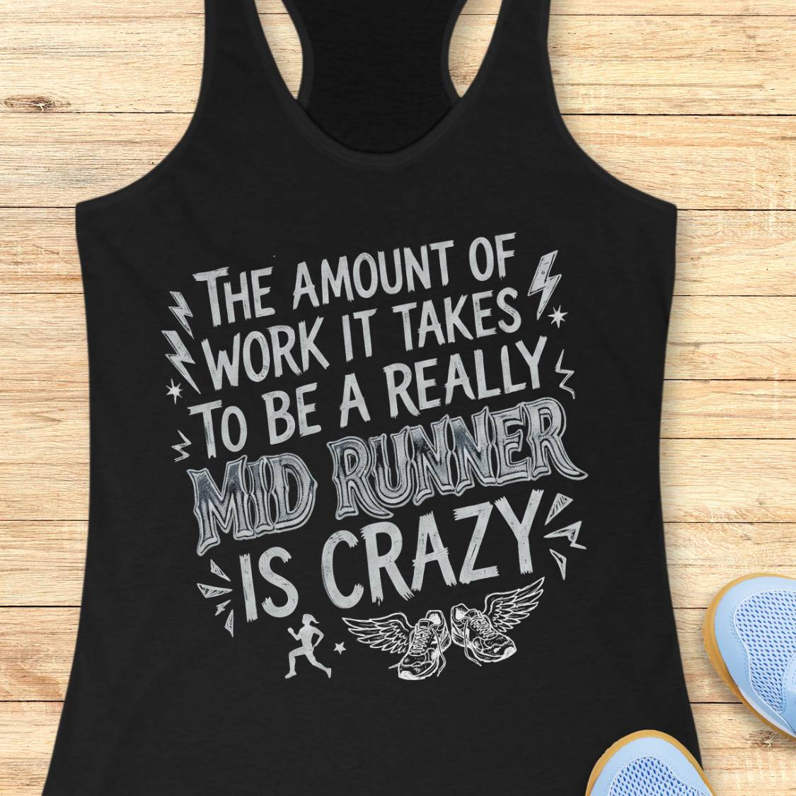 A black racerback tank top with a white graphic text print reading 'THE AMOUNT OF WORK IT TAKES TO BE A REALLY MID RUNNER IS CRAZY' laid flat on a light wood background, with partial blue sneakers visible at the bottom right.
