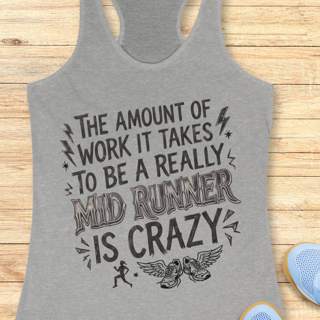 A heather gray racerback tank top with a black graphic text print that reads 'THE AMOUNT OF WORK IT TAKES TO BE A REALLY MID RUNNER IS CRAZY' with small running and winged shoe illustrations, shown on a light wood background with partial blue shoes visible at the edges.