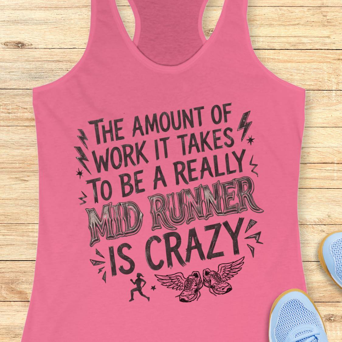 A pink racerback tank top with a black graphic text that reads 'THE AMOUNT OF WORK IT TAKES TO BE A REALLY MID RUNNER IS CRAZY' with lightning bolts, stars, a running figure, and winged shoes, shown on a light wood background with partial blue sneakers at the edges.