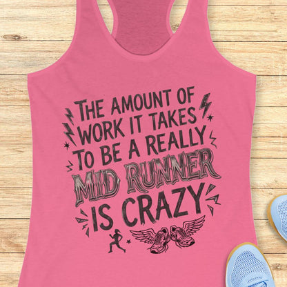 A pink racerback tank top with a black graphic text that reads 'THE AMOUNT OF WORK IT TAKES TO BE A REALLY MID RUNNER IS CRAZY' with lightning bolts, stars, a running figure, and winged shoes, shown on a light wood background with partial blue sneakers at the edges.