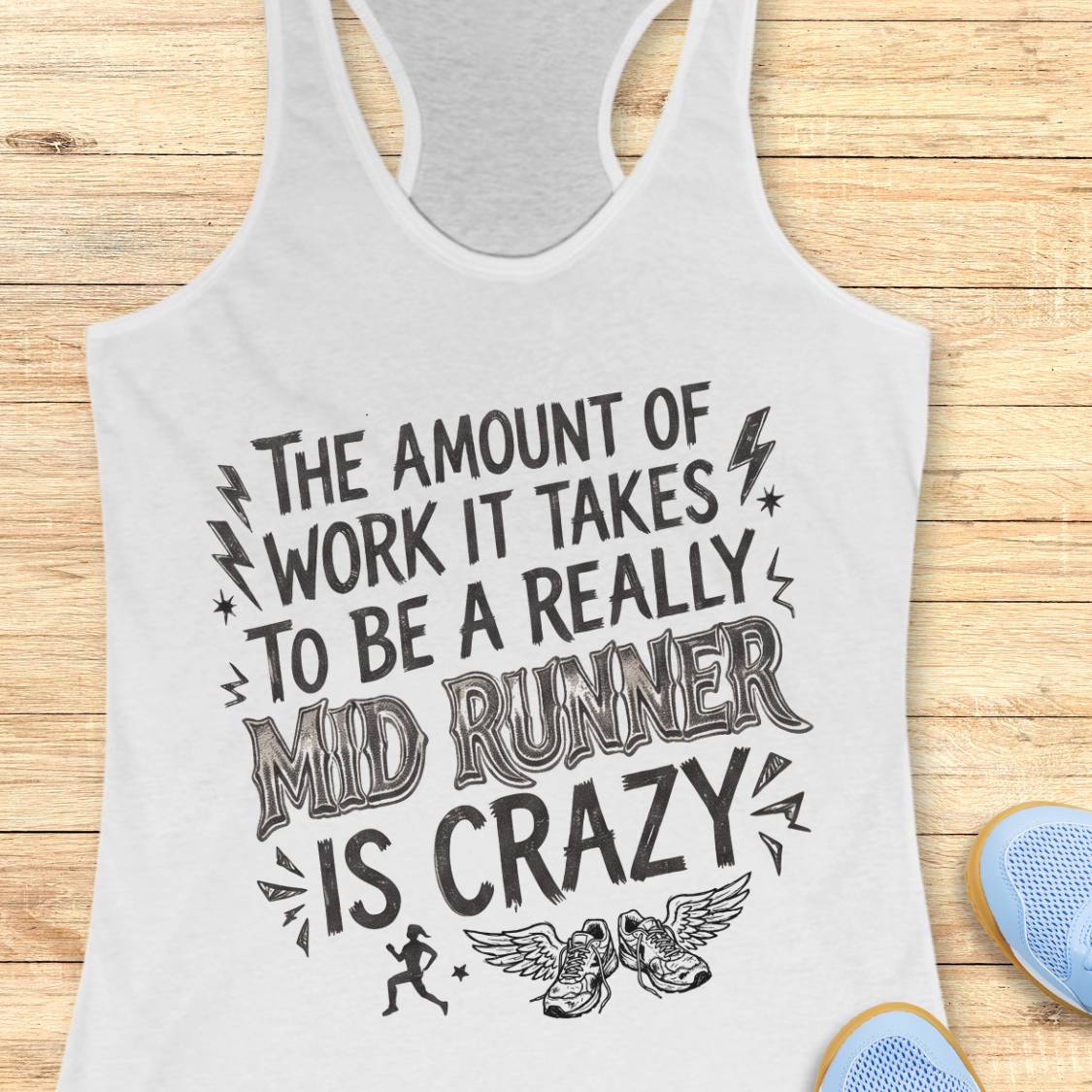 A white racerback tank top with a black graphic text print reading 'THE AMOUNT OF WORK IT TAKES TO BE A REALLY MID RUNNER IS CRAZY' with small running, winged shoe, and lightning accents, shown on a wood plank background with partial blue sneakers at the edges.
