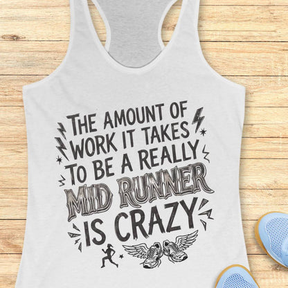 A white racerback tank top with a black graphic text print reading 'THE AMOUNT OF WORK IT TAKES TO BE A REALLY MID RUNNER IS CRAZY' with small running, winged shoe, and lightning accents, shown on a wood plank background with partial blue sneakers at the edges.