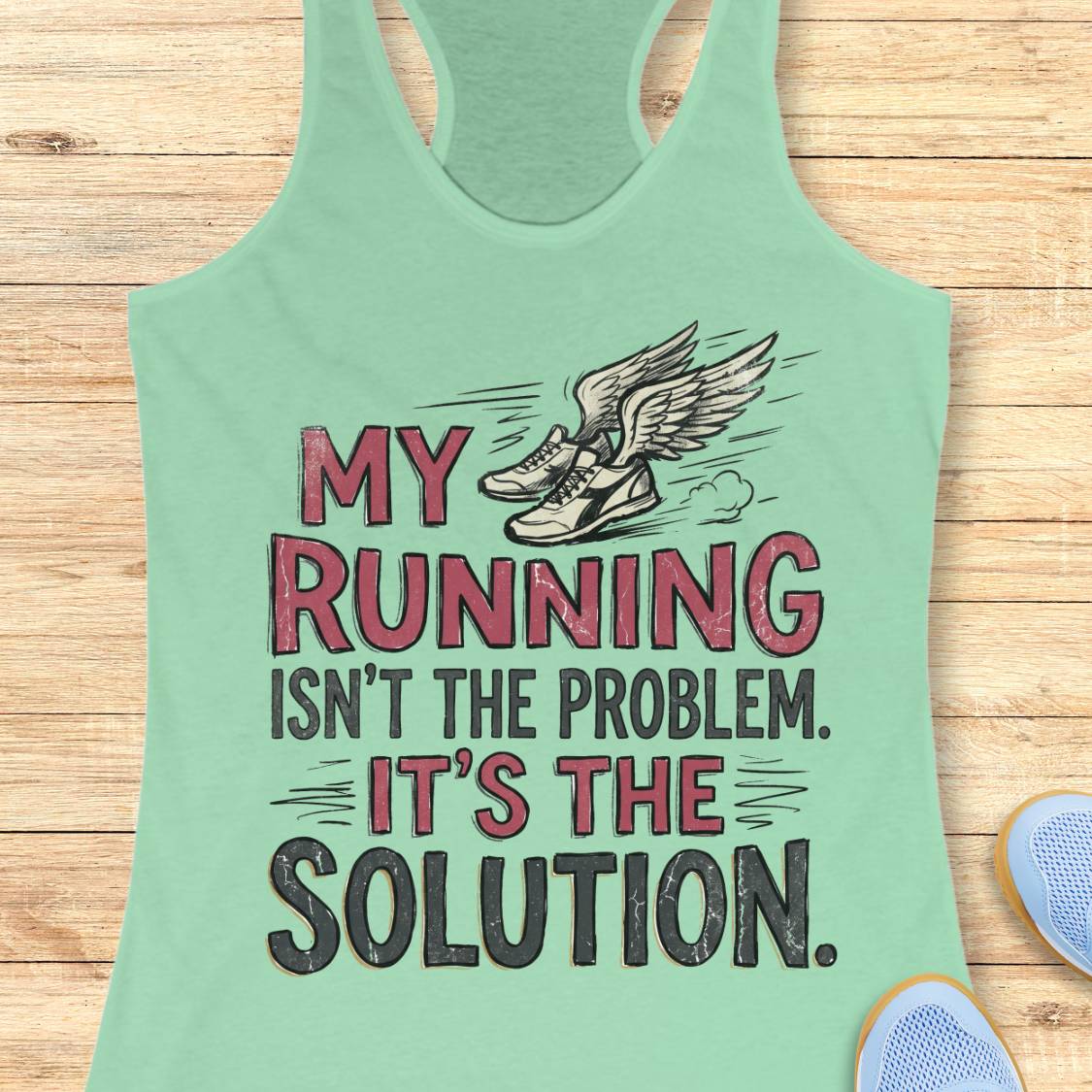 A mint green racerback tank top laid flat on a light wood background, featuring a graphic print of winged running shoes and the text 'MY RUNNING ISN'T THE PROBLEM. IT'S THE SOLUTION.