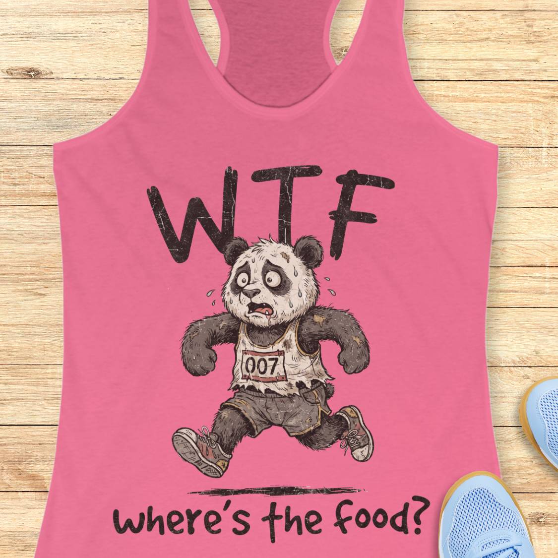 A pink racerback tank top laid flat on a light wood surface, featuring a cartoon panda runner with the text 'WTF' above and 'where's the food?' below. Partial blue shoes are visible at the edges as styling props.