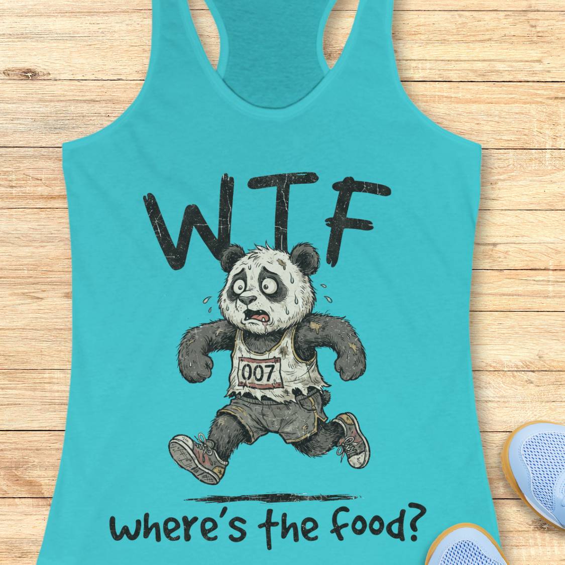 A turquoise racerback tank top laid flat on a light wood surface, featuring a distressed black graphic of a panda running with the text 'WTF' above and 'where's the food?' below. A partial blue shoe is visible at the bottom right.