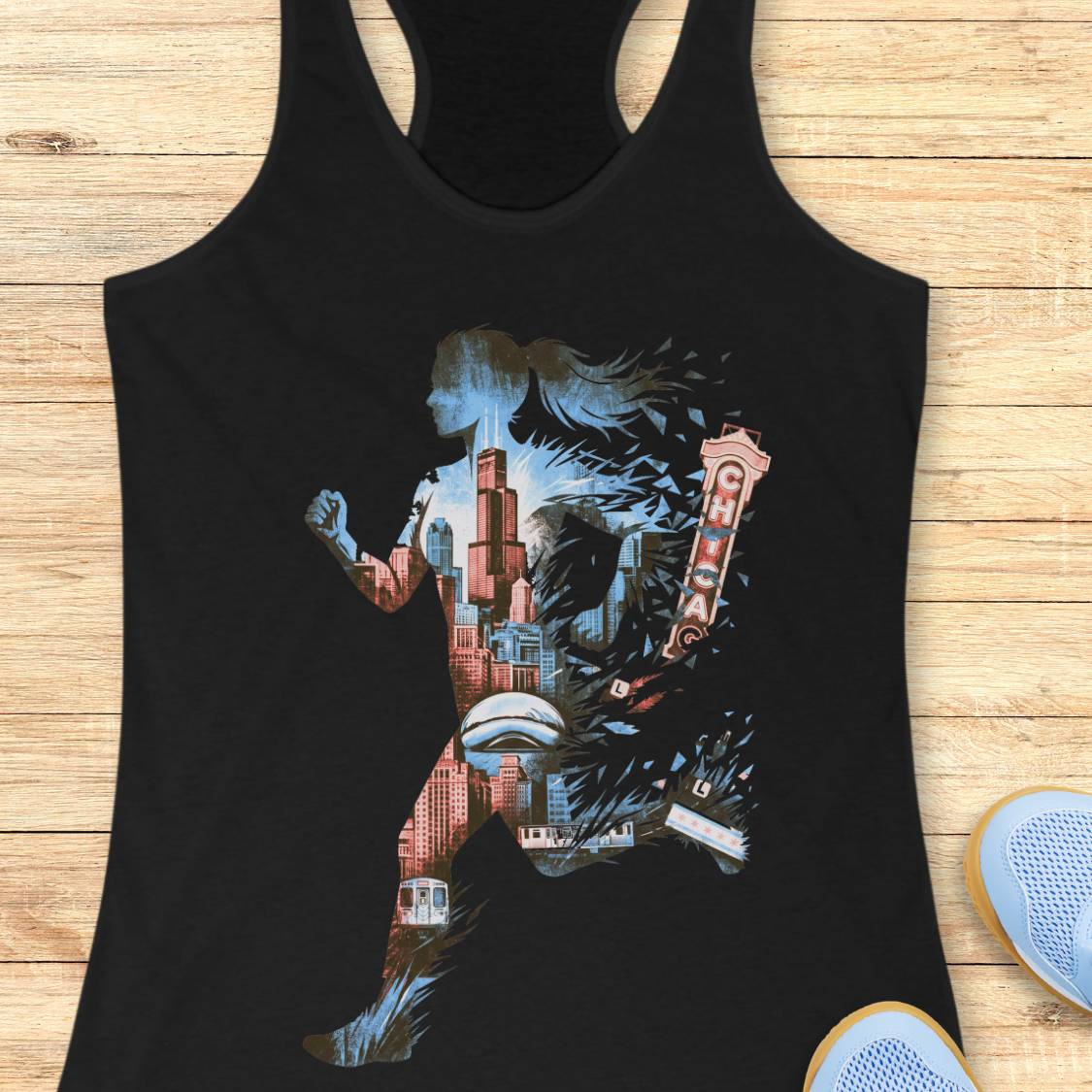 A black racerback tank top laid flat on a light wood surface, featuring a large multicolor graphic of a running figure filled with Chicago cityscape elements and the Chicago skyline.