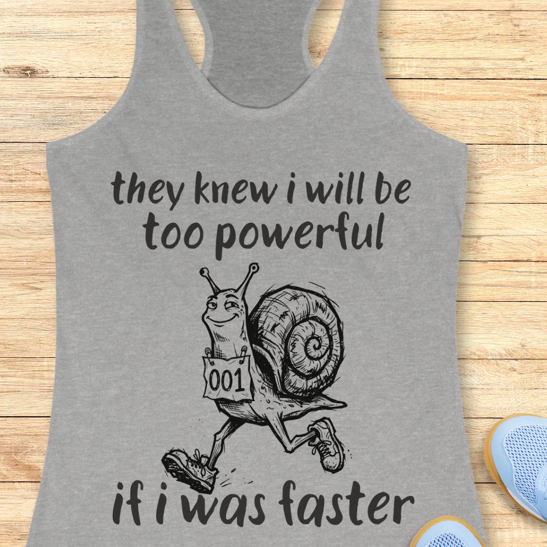 A heather gray racerback tank top laid flat on a light wood surface, featuring a black graphic of a snail running with the text 'they knew i will be too powerful if i was faster.' A partial blue shoe is visible at the lower right edge.