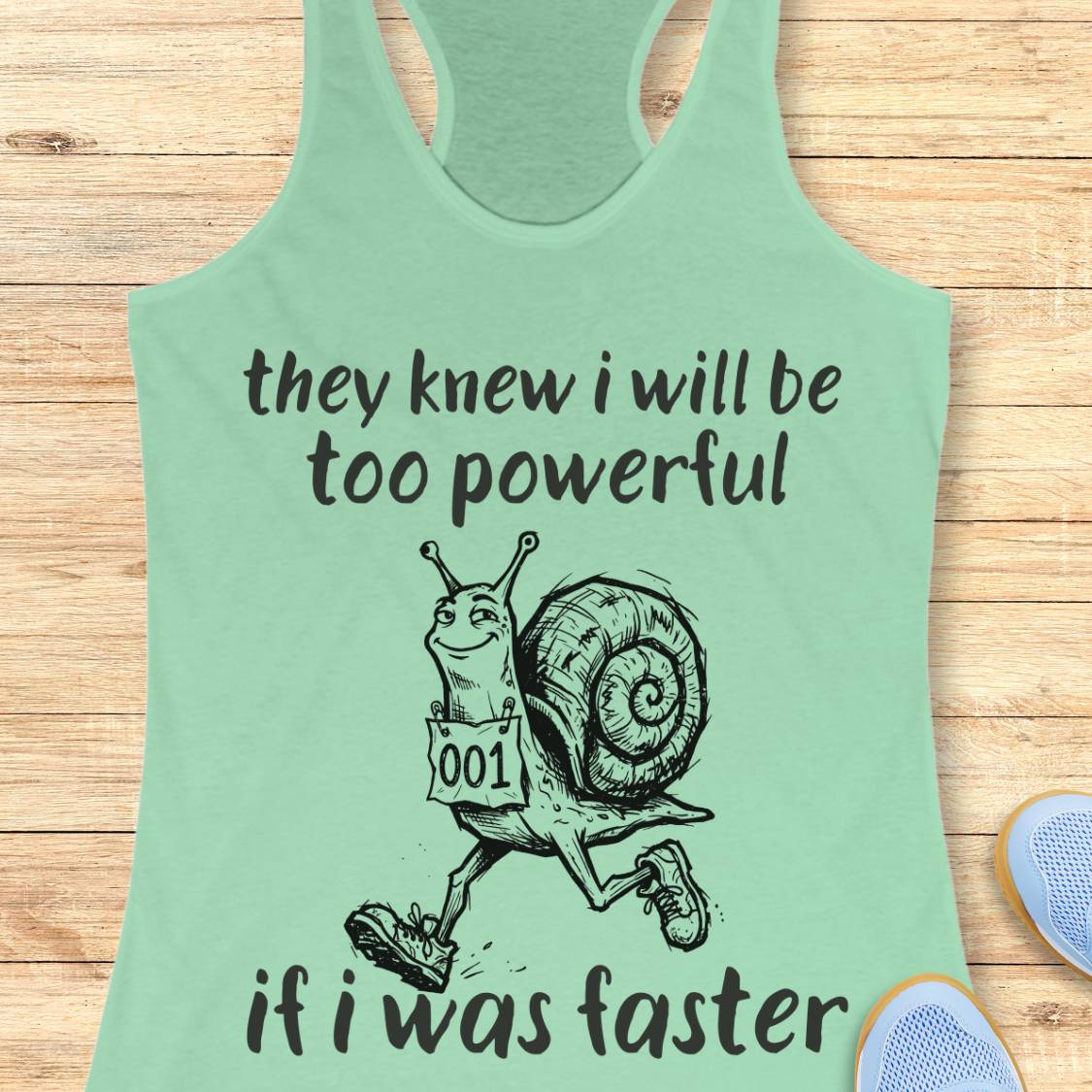 A mint green racerback tank top laid flat on a light wood background, featuring a black graphic of a cartoon snail running with the text 'they knew i will be too powerful if i was faster'.
