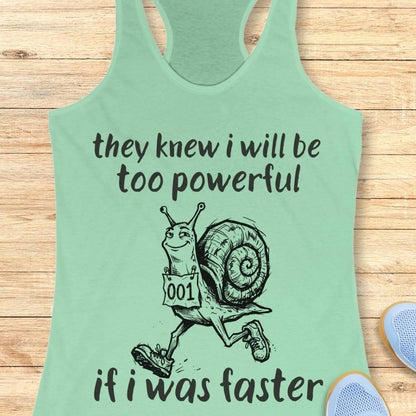 A mint green racerback tank top laid flat on a light wood background, featuring a black graphic of a cartoon snail running with the text 'they knew i will be too powerful if i was faster'.