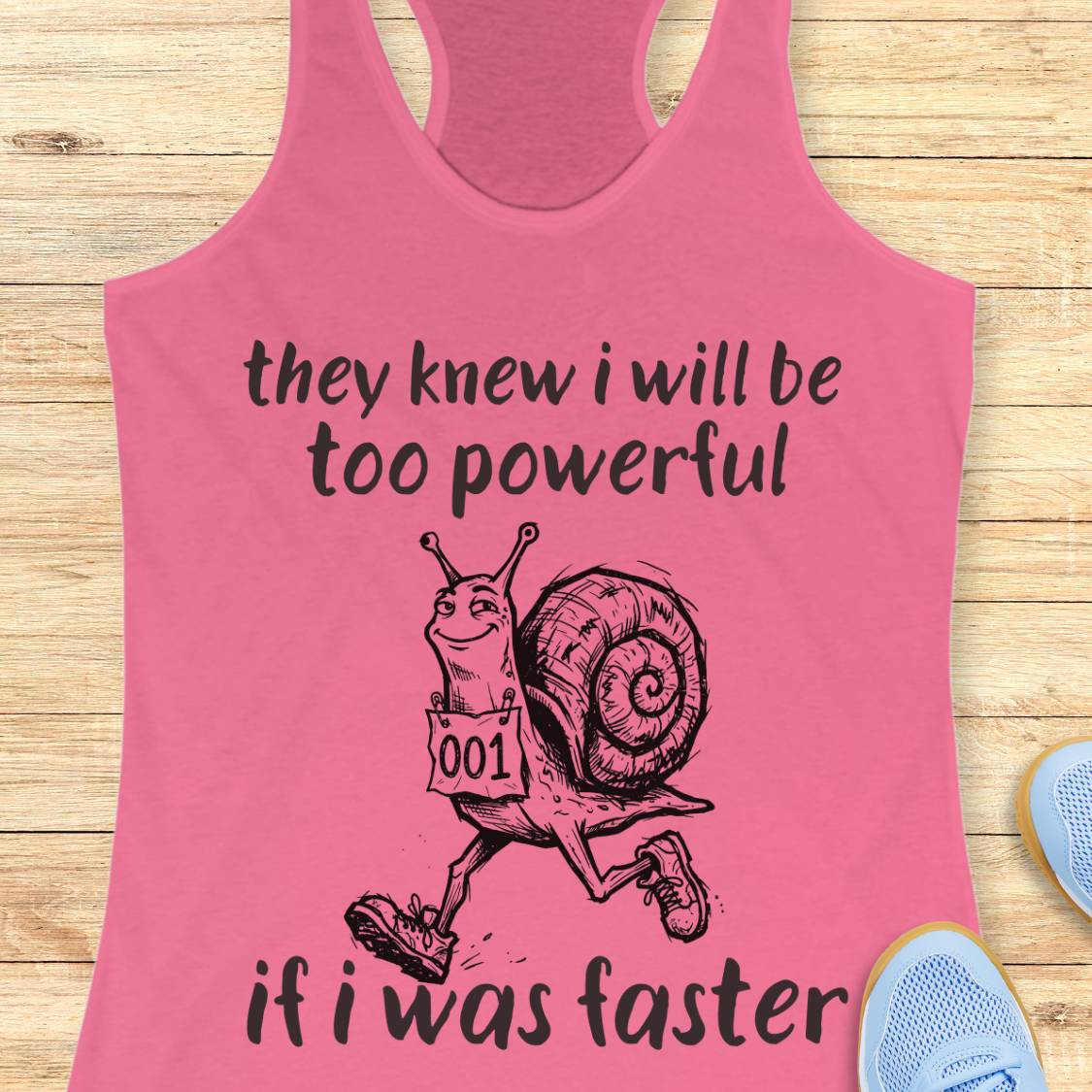 A pink racerback tank top laid flat on a light wood surface, featuring a black graphic of a snail running with the text 'they knew i will be too powerful if i was faster.' Partial blue shoes are visible at the edges as styling props.