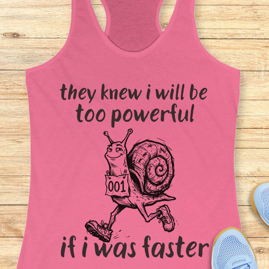 A pink racerback tank top laid flat on a light wood surface, featuring a black graphic of a snail running with the text 'they knew i will be too powerful if i was faster.' Partial blue shoes are visible at the edges as styling props.
