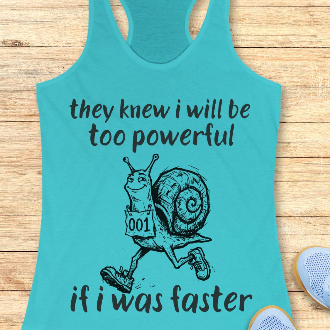A turquoise racerback tank top laid flat on a light wood surface, featuring a black graphic of a snail running with the text 'they knew i will be too powerful if i was faster.' A partial white shoe is visible at the lower right edge.