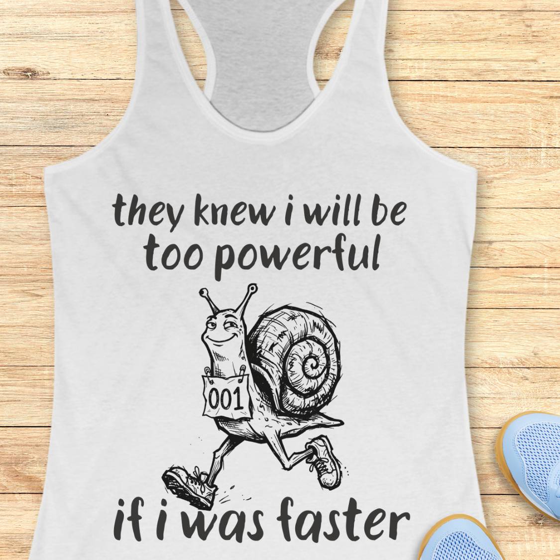 A white racerback tank top laid flat on a light wood surface, featuring a black graphic of a snail running with the text 'they knew i will be too powerful if i was faster.' Partial blue shoes are visible at the bottom right as styling props.