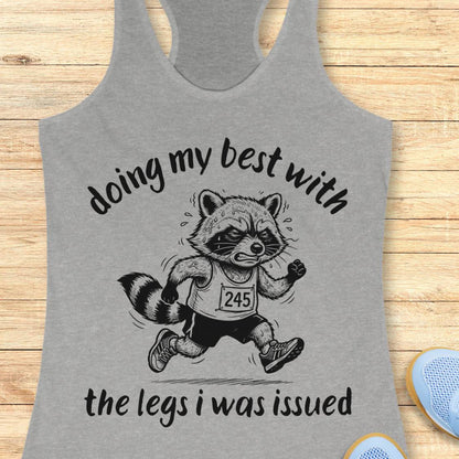 A heather gray racerback tank top laid flat on a light wood surface, featuring a black graphic of a raccoon runner with the text 'doing my best with the legs i was issued'.