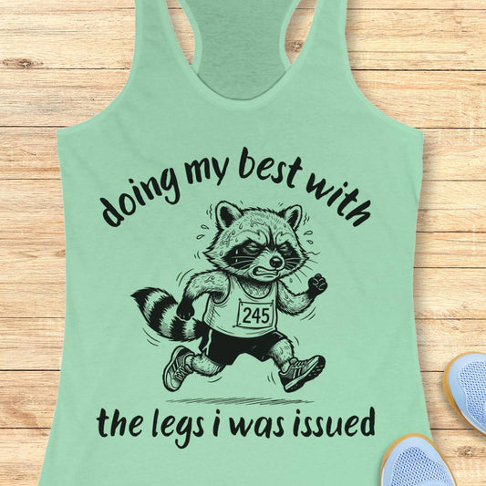 A mint green racerback tank top laid flat on a light wood background, featuring a black graphic of a raccoon running with the text 'doing my best with the legs i was issued'.