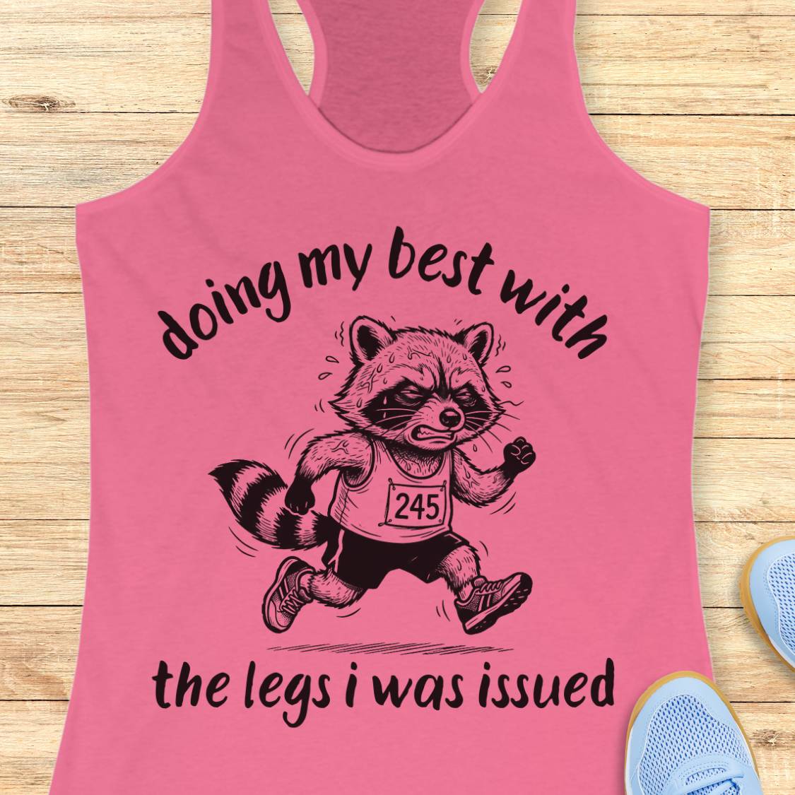 A pink racerback tank top laid flat on a light wood surface, featuring a black graphic of a raccoon running with the text 'doing my best with the legs i was issued'. Partial blue shoes are visible at the bottom right as styling props.