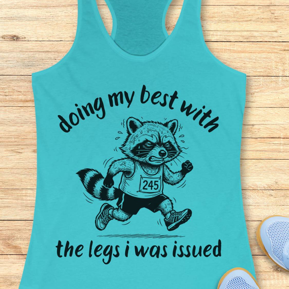A turquoise racerback tank top laid flat on a light wood surface, featuring a black graphic of a raccoon running with the text 'doing my best with the legs i was issued'. A partial white shoe is visible at the bottom right.