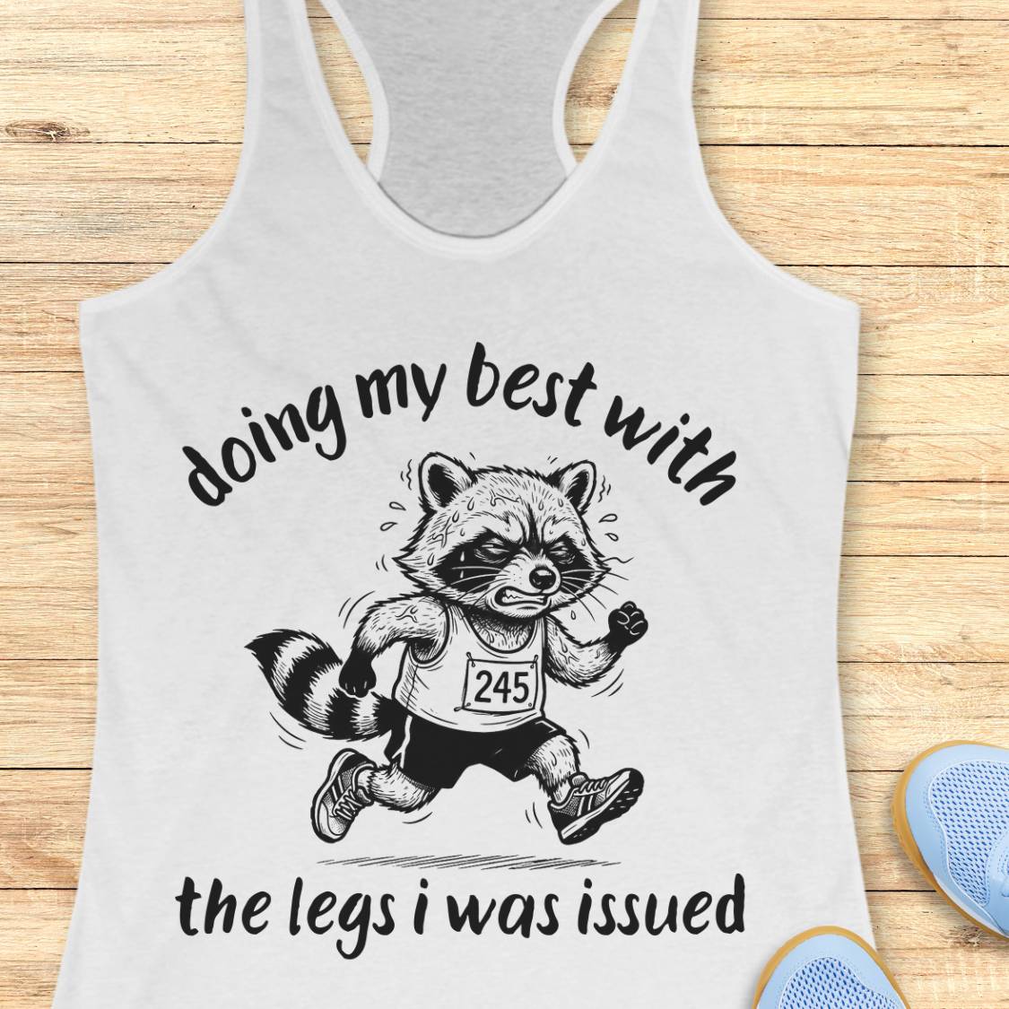 A white racerback tank top laid flat on a light wood surface, featuring a black graphic of a raccoon runner with the text 'doing my best with the legs i was issued'. Partial blue shoes are visible at the bottom right as styling props.