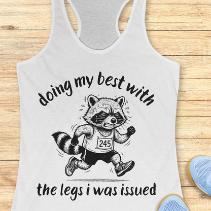 A white racerback tank top laid flat on a light wood surface, featuring a black graphic of a raccoon runner with the text 'doing my best with the legs i was issued'. Partial blue shoes are visible at the bottom right as styling props.