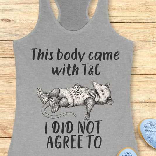 A heather gray racerback tank top laid flat on a light wood background, featuring a black graphic of a sleeping raccoon wearing a race bib with the text 'This body came with T&C I DID NOT AGREE TO'.