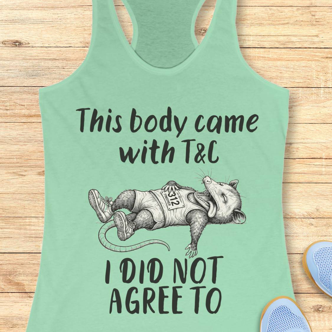 A mint green racerback tank top laid flat on a light wood background, featuring a black graphic of a sleeping opossum wearing a shirt that says 'T&C' and the text 'This body came with T&C I DID NOT AGREE TO'. Partial blue shoes are visible at the bottom right.