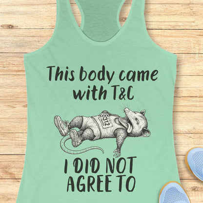 A mint green racerback tank top laid flat on a light wood background, featuring a black graphic of a sleeping opossum wearing a shirt that says 'T&C' and the text 'This body came with T&C I DID NOT AGREE TO'. Partial blue shoes are visible at the bottom right.