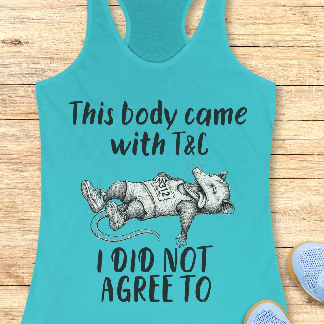 A turquoise racerback tank top laid flat on a light wood surface, featuring a black graphic of a sleeping raccoon wearing a vest with 'RACER 252' and the text 'This body came with T&C I DID NOT AGREE TO'. Partial blue shoes are visible at the bottom right edge.